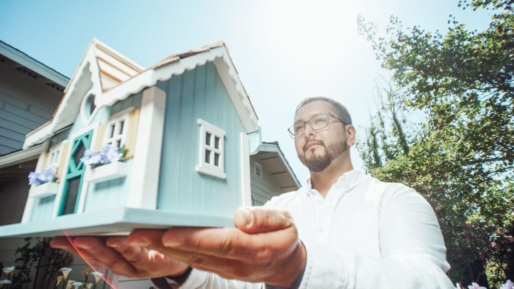 pexels-photo-7578936 A man holds a miniature house model outdoors on a sunny day, symbolizing home buying.