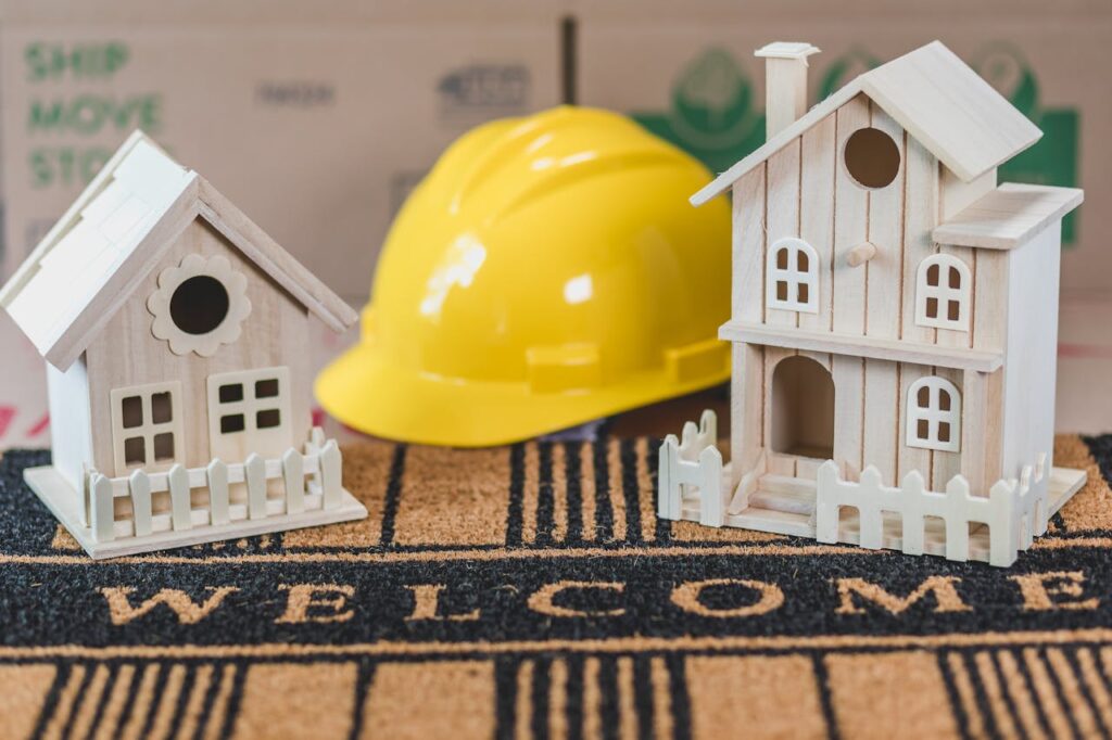 pexels-photo-8293708 Miniature wooden houses on a welcome mat with a yellow hard hat in the background, symbolic of home building or construction.